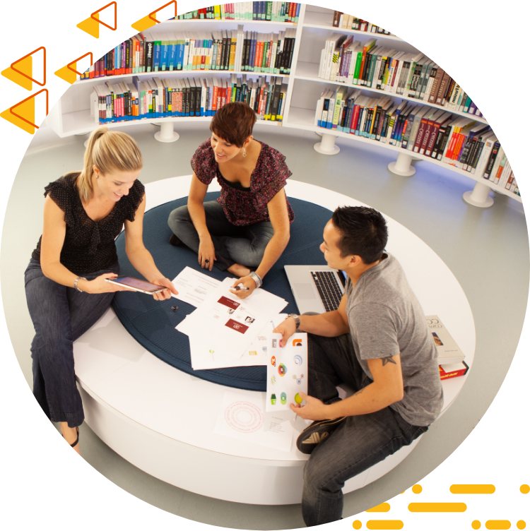 Aerial photo of three students collaborating on design work while seated on a large ottoman, 书籍是背景.